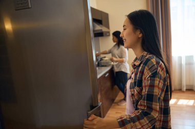 Yetişkin bir kadın tavada yemek pişirirken gülümseyen genç bir kadının buzdolabının kapısını açması.