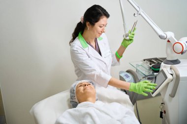 Kozmetik bilimci modern ekipman, kep ve kabarık bornoz giymiş bir hasta kullanarak bir donanım kozmetoloji prosedürü uyguluyor.