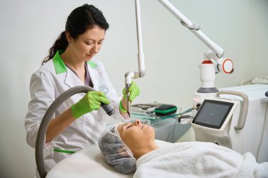 Kozmetik kliniğindeki modern donanım kozmetolojisinin yenilenme prosedürü, kep ve kabarık bornoz giymiş bir hasta