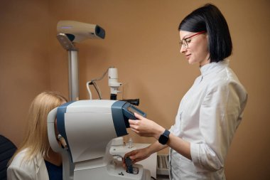 Genç bir göz doktoru çalışmalarında özel ekipman kullanır, genç bir kadına bir uzman tarafından bakılır.