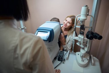 Sarışın ve küçük kızı otomatik fraktometre başında oturuyorlar. Bir uzman kızların gözlerini kontrol ediyor.