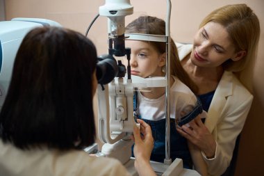 Specialist checks the vision of a young patient using modern equipment, the girl sits in her mother arms clipart