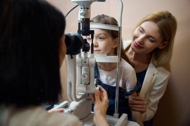 Female ophthalmologist checks the vision of a young patient using modern equipment, the girl sits in her mothers arms clipart