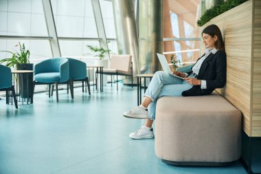 Elinde dizüstü bilgisayarı olan genç bir bayan bir sedirin üzerinde otururken çalışıyor.