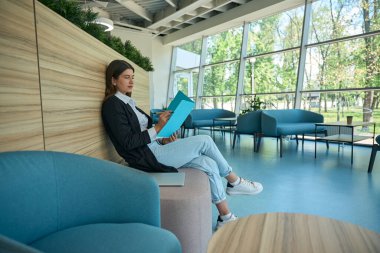 Ceketli beyaz kadın elinde mavi bir klasörle oturuyor.