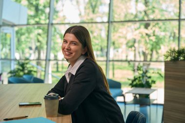 Genç çalışan masada ceketle oturuyor ve kameraya gülümsüyor.