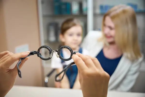 stock image Mom and little daughter are sitting at the table at doctors appointment, ophthalmologist is holding diagnostic glasses in his hands