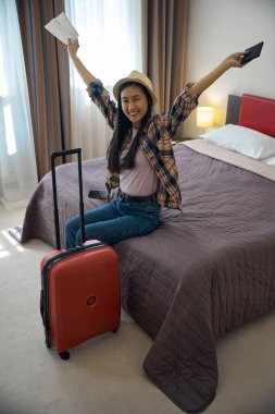 Otel odasında rahat bir yatakta oturan mutlu bir yolcu. Elinde pasaport ve uçuş kartı var.