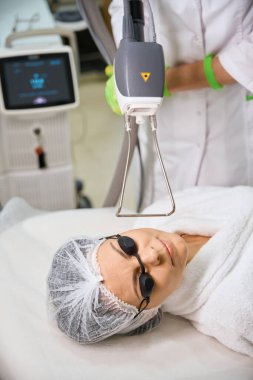 Dermatolog işinde modern bir lazer cihazı kullanıyor, hasta güvenlik gözlüğü ve şapka takıyor.