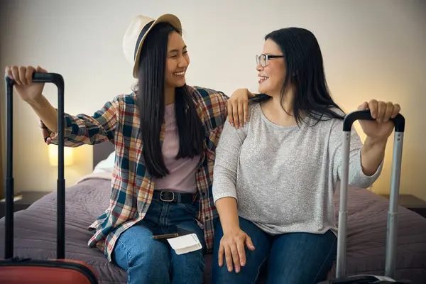 stock image Two joyful Asian travelers with travel bags seated on comfortable bed in suite looking at each other