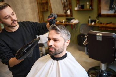 Yüksek kalifiye saç stilisti yakışıklı yetişkin erkek alnından özel fırça ve saç kurutma makinesi kullanıyor, havalı saç stili yapıyor, berber servisi yapıyor.