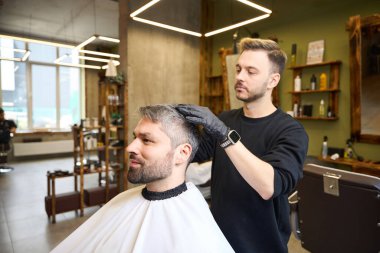 Tecrübeli saç stilisti, berber dükkanını ziyaret eden, saç modelini krem kullanarak modelleyen yetişkin, yakışıklı adamın şık saç stiline son rötuşları yapıyor.