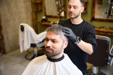 Deneyimli berber, yetişkin müşterilere masaj yapıyor saç kesiminden sonra saçlarını modelliyor ve şekillendiriyor. Kalıp merhemi kullanıyor, müşteriye aynada bakıyor.