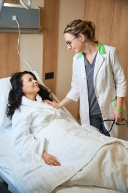 Klinik çalışanı koğuştaki bir hastayla iletişim kuruyor. Doktorun elinde steteskop var.