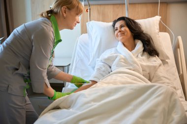 Gülümseyen hemşire, beyaz kabarık bornozlu kadın bir kadına serum takıyor.