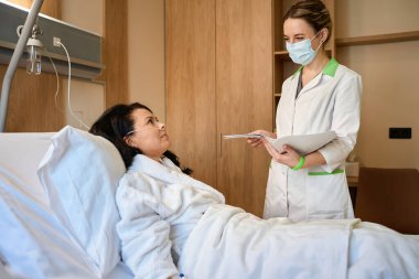 Koruyucu maskeli kadın doktor hastane odasında bir bayanla iletişim kuruyor, kadın rahat bir yatakta yatıyor.
