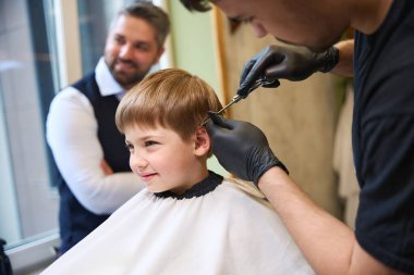 Modern berber dükkanının deneyimli ustası. Erkek kulaklarının üstündeki saçı kesmek için keskin bir makas kullanıyor. Küçük müşteriye şık bir saç stili yapıyor.