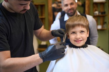 High qualified barber cutting excess hair above little client ears who sitting on the barber chair above the cope, making boy look more stylish and mature clipart