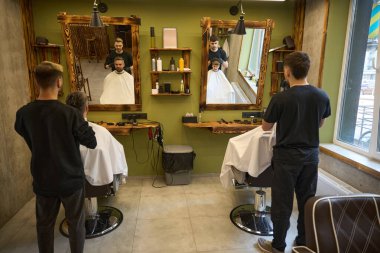 İki berber, berber dükkanlarını ziyaret eden yetişkin bir adam ve küçük bir çocuğa saç kesimi yaptırıyor. Müşterilerin duvardaki büyük aynalara bakıyorlar.
