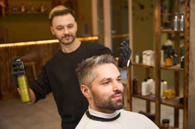 Gülümseyen berber saç spreyi sıkar yakışıklı adama saç kesimi, stil sahibi görünümü bitirir, müşteri saç stilinden tatmin olur ve kişiliğini yansıtır.