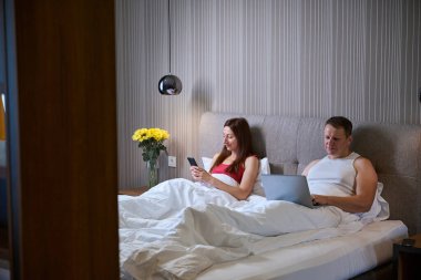 Yatakta, modern aletleri olan, yanındaki komodinin üzerinde sarı çiçekleri olan evli bir çift.