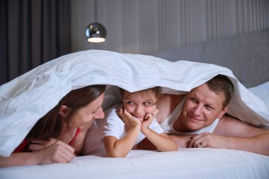 Anne ve baba, büyük bir yatakta battaniyeden yapılmış bir kulübede küçük bir kızla neşeyle gülümsüyorlar.