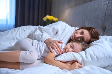Küçük kızı ve annesi büyük, rahat bir yatakta uzanıyorlar, çocuk tatlı tatlı kestiriyor.