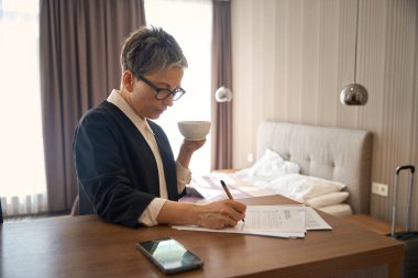 Gözlüklü şık yaşlı kadın formu dolduruyor, elinde çay tutuyor, otel odasında dikiliyor. Seyahat, iş konsepti