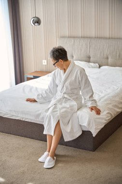 Beyaz bornozlu gözlüklü yetişkin bir kadın otel odasında yatakta oturuyor. Seyahat, insanlar, yaşam tarzı konsepti