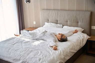 Beyaz bornozlu, gözlüklü, otel odasında yatakta dinlenen gülümseyen olgun bir kadın. Seyahat, insanlar, yaşam tarzı konsepti