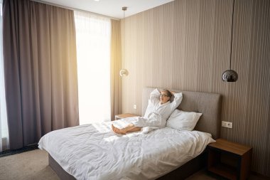 Beyaz bornozlu, otel odasında yatakta dinlenen mutlu yaşlı bayan. Seyahat, insanlar, yaşam tarzı konsepti