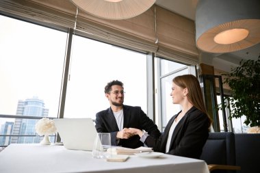 Mutlu iş ortakları restoranda otururken anlaşmadan sonra el sıkışıyor, dizüstü bilgisayar masada. İş, ortaklık, teknoloji kavramı