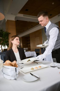 Garson elinde iki tabak var, restoranda bir kadın ona bakarken masaya servis yapıyor. Gurme hazırlığı, servis kavramı