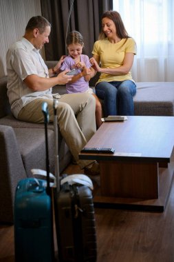 Ebeveynler ve kızları rahat bir koltukta oturuyorlar, baba kıza cep telefonundan bir şey gösteriyor.