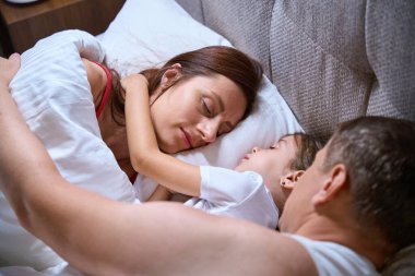 Anne, baba ve küçük kız büyük bir yatakta uyukluyorlar, aileler kıza sarılıyorlar.