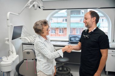 Uzman, el sıkışan yaşlı bir bayanı selamlıyor, dişçi kliniğinde modern ekipmanlar var.