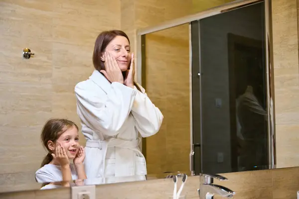 stock image Young woman and a little girl wash themselves in front of a mirror, in a modern design bathroom