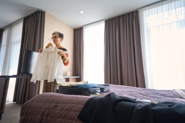 Otel odasında bavulları açarken elinde beyaz gömlek tutan gözlüklü yetişkin bir kadın. Seyahat kavramı