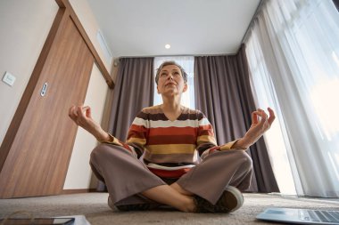 Son sınıf öğrencisi, otel odasında dizüstü bilgisayarla lotus pozisyonunda oturmuş yoga yapıyor. Seyahat, iş konsepti