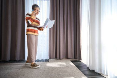 Gözlüklü olgun bir iş kadını akıllı telefondan konuşuyor ve elinde belgeler, dizüstü bilgisayar otel odasında seyahat, iş konsepti...