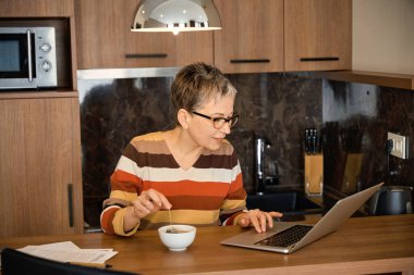 Kadehler içinde çayını yudumlayan ve dizüstü bilgisayarında oteldeki masada oturan yaşlı bir kadın. Seyahat, iş konsepti
