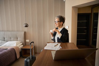 Otel odasında dizüstü bilgisayar ve kağıtlarla çalışırken çay içen güzel bir iş kadını. Seyahat, iş konsepti