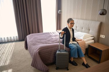 Otel odasında gözlüklü, elinde bavuluyla yatakta oturan şık bir kadın. Seyahat, insanlar, yaşam tarzı konsepti
