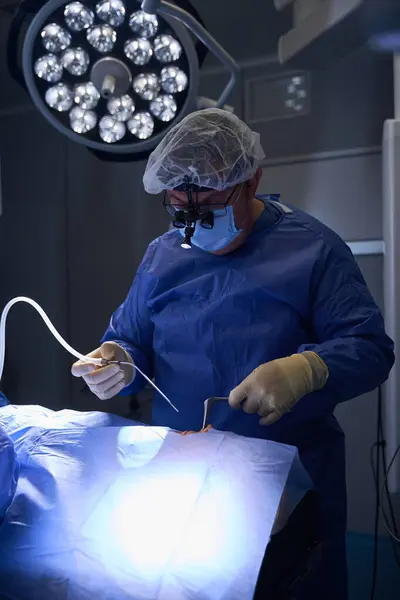 stock image Surgeon in blue surgical attire is focused on performing a procedure under bright, overhead operating room lights