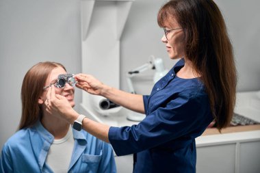 Göz doktoru, hastanın yanında duruyor. Göz muayenesi için deneme çerçevesini ayarlıyor.