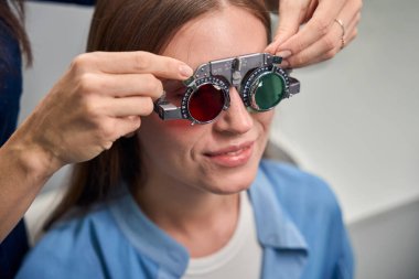 Yetişkin beyaz bir hasta, göz muayenesi sırasında göz doktorunun rehberliğinde çeşitli lenslerle bir deneme çerçevesi takıyor.