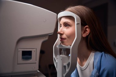 Genç bir kadın, göz taraması için klinik bir ortamda detaylı bir göz taraması hazırlayan göz sağlığı cihazının önünde oturuyor.