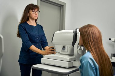 Kadın optometrist, optalmik cihazın önündeki hastayı test etmek için uygun hizayı sağlayarak konumlandırır.