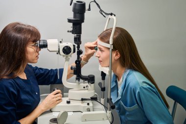 Yetişkin kadın optometrist, klinik bir ayarda kesik lamba kullanarak hastanın gözlerini dikkatlice inceliyor.