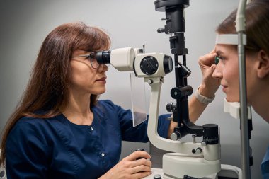 Göz muayenesi yapan kadın optometrist yarık lamba kullanarak hastaların gözlerini dikkatle inceliyor.